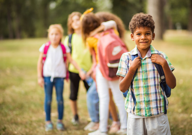 one kid posing with backpack outdoors with a group of kids behind him